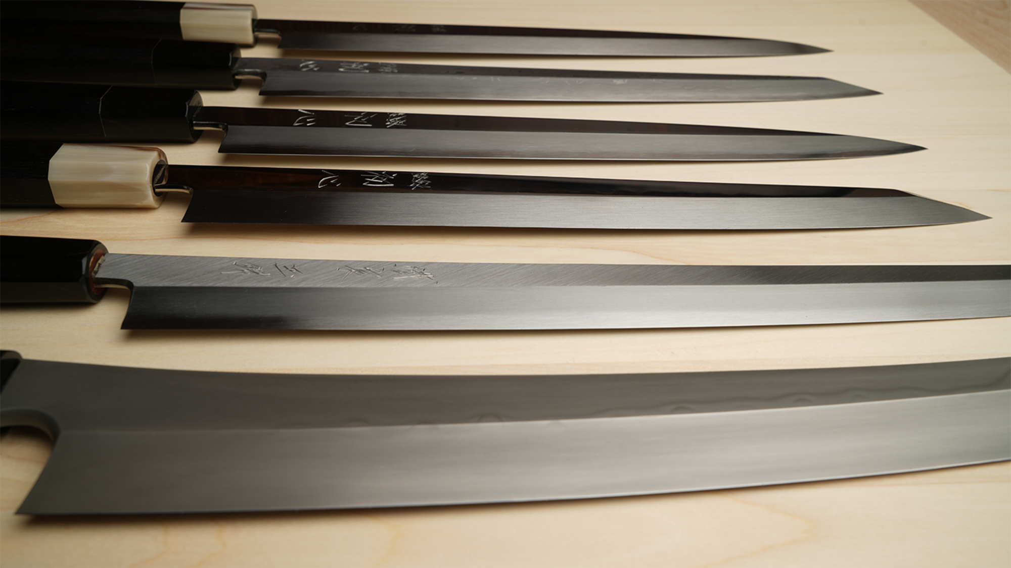 A set of Japanese yanagi knives with long  steel blades and black handles, arranged horizontally on a light wood surface.