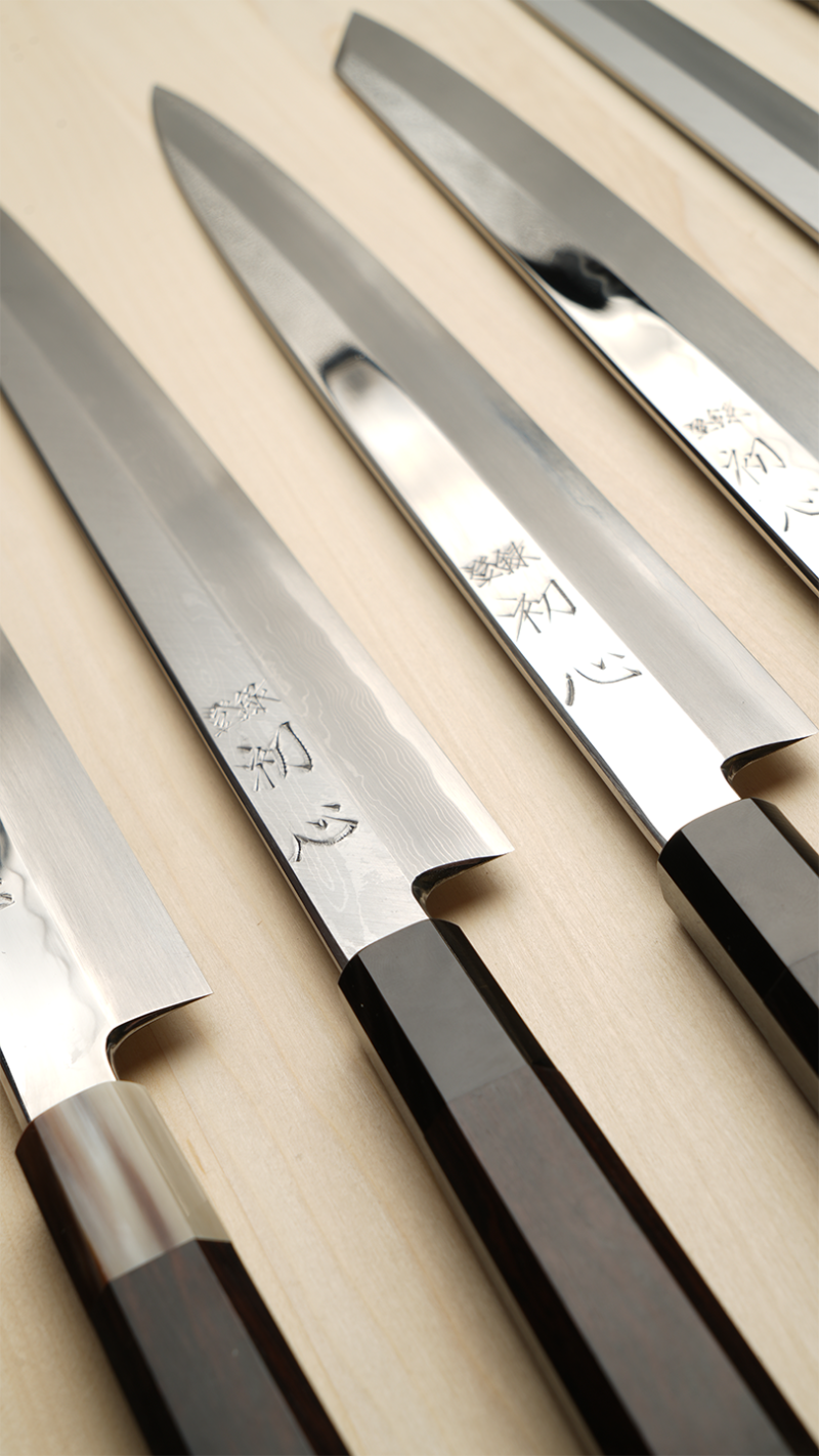Japanese Yanagi knives with polished steel blades and black handles, arranged diagonally on a light beige wooden surface. Each blade features engraved Japanese characters.
